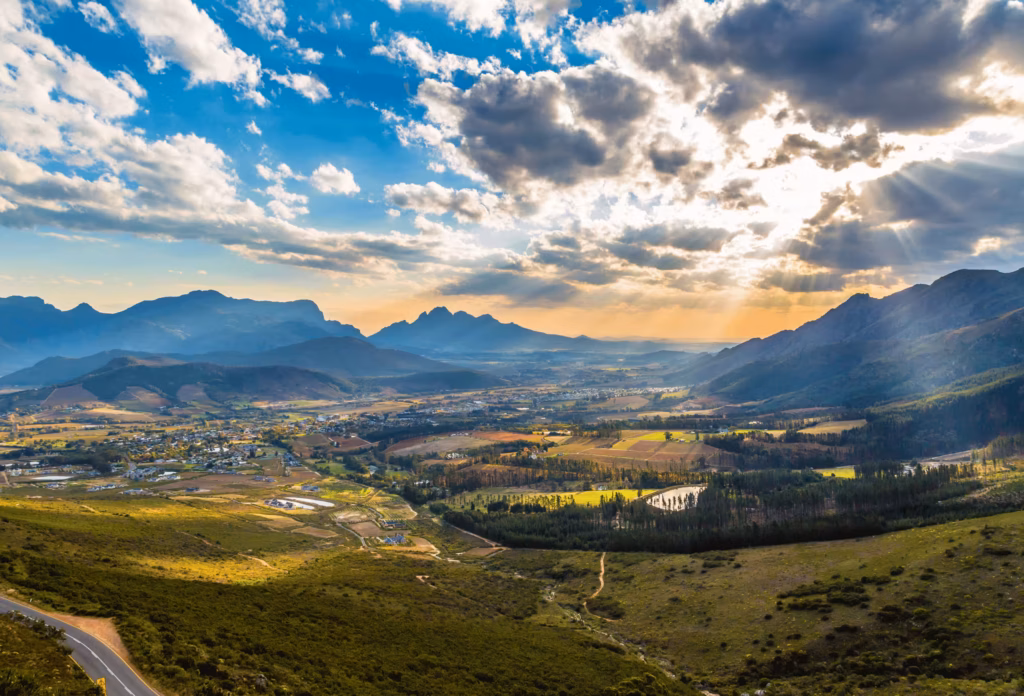 Landskapet i Franshhoek i Sydafrika sett uppifrån, med berg i bakgrunden och solen som lyser igenom mörkare moln